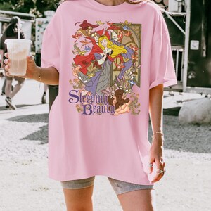 May include: A light pink short-sleeve t-shirt featuring a graphic print of the character Aurora from Sleeping Beauty, surrounded by forest animals and birds. The text Sleeping Beauty is printed in a purple serif font at the bottom of the design.