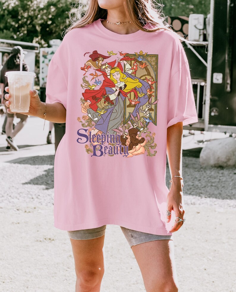 May include: A light pink short-sleeve t-shirt featuring a graphic print of the character Aurora from Sleeping Beauty, surrounded by forest animals and birds. The text Sleeping Beauty is printed in a purple serif font at the bottom of the design.