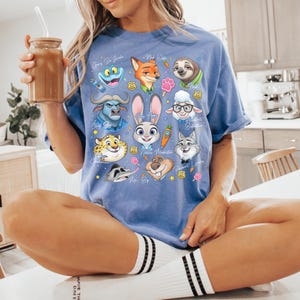 Puede incluir: Una camiseta de manga corta en color azul b&iacute;garo con un estampado gr&aacute;fico colorido de varios personajes animales animados de Zootopia, incluidos Judy Hopps, Nick Wilde y Flash, dispuestos en un collage con peque&ntilde;os iconos como zanahorias.