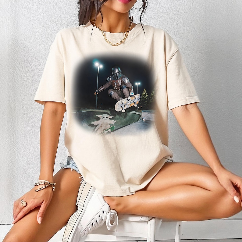 May include: Beige t-shirt featuring a graphic of The Mandalorian skateboarding in a skate park at night. A cartoon version of Grogu is also on a skateboard. The shirt is a casual, unisex style.