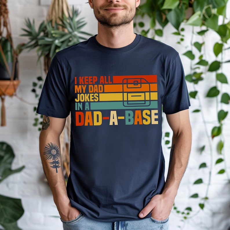 May include: Navy blue t-shirt with the text "I KEEP ALL MY DAD JOKES IN A DAD-A-BASE" in a retro color scheme. The design includes a graphic of a floppy disk. A casual tee for a dad.