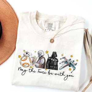 Puede incluir: Camiseta color crema con personajes de Star Wars: BB-8, Grogu, The Mandalorian, Darth Vader y un caza X-wing. La frase "May the Force be with you" está escrita en guion negro. La camiseta tiene cuello redondo.