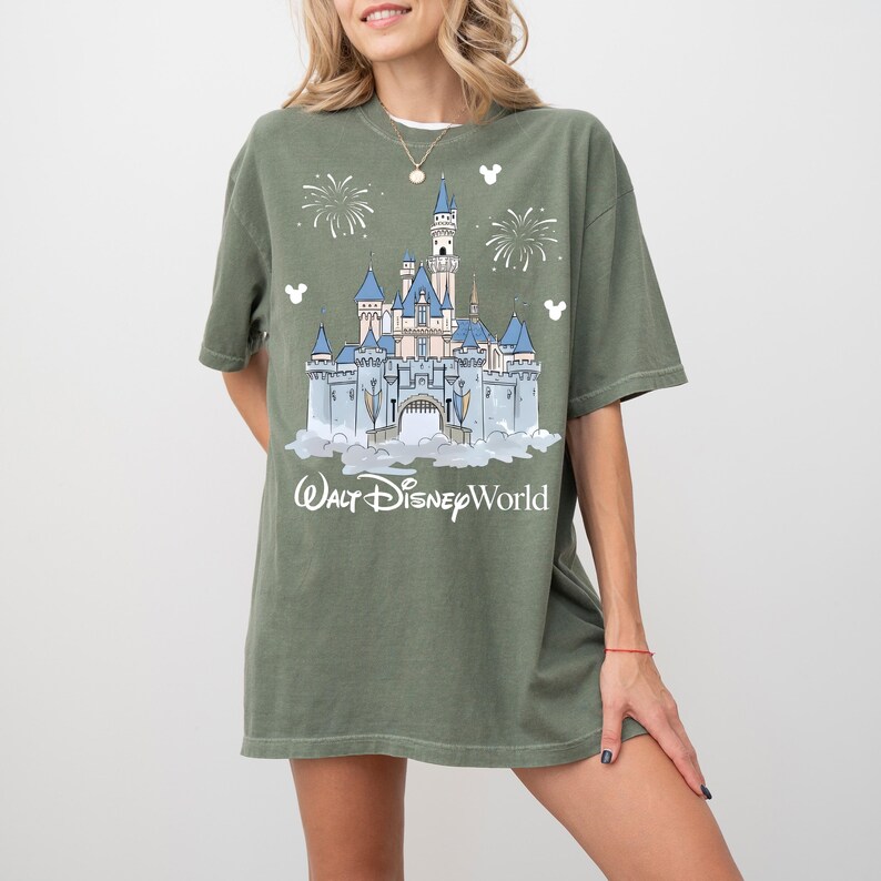 May include: Olive green t-shirt with a graphic of Cinderella's castle, blue turrets, and fireworks. The text "Walt Disney World" is printed below the castle in white. The shirt has short sleeves and a relaxed fit.