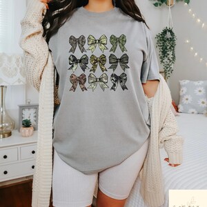May include: A gray t-shirt featuring nine bow designs in various patterns and colors, including green, brown, and black. The bows are arranged in a grid pattern. The shirt is part of a casual clothing collection.
