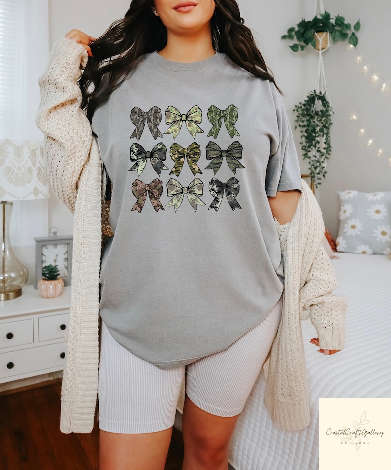 May include: A gray t-shirt featuring nine bow designs in various patterns and colors, including green, brown, and black. The bows are arranged in a grid pattern. The shirt is part of a casual clothing collection.