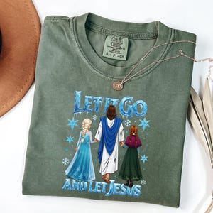May include: An olive green t-shirt with the phrase "Let It Go and Let Jesus" in blue and white. The graphic features illustrations of Jesus, Elsa, and Anna. A gold necklace and a brown hat are also visible.