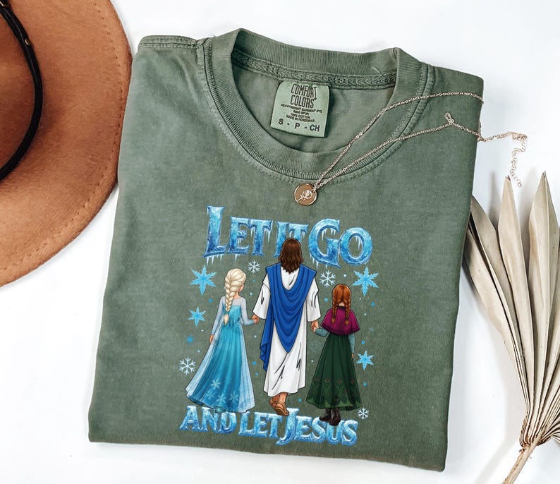 May include: An olive green t-shirt with the phrase "Let It Go and Let Jesus" in blue and white. The graphic features illustrations of Jesus, Elsa, and Anna. A gold necklace and a brown hat are also visible.