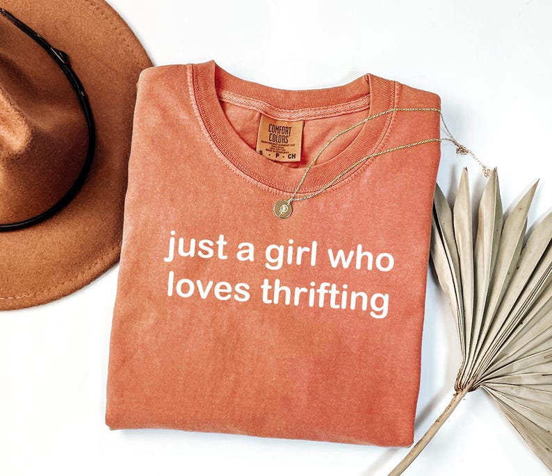 May include: Coral-colored t-shirt with the text "just a girl who loves thrifting" in white. A gold necklace with a pendant and a brown hat are also in the image. The shirt has a crew neck and a tag that says "Comfort Colors".