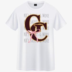 May include: White t-shirt featuring a large maroon letter "G" with a gold outline and a pink bow. The shirt also has the text "KK'S SHIRTS & MORE" in a repeating pattern. A simple, casual design.