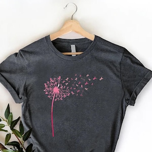 May include: Dark gray t-shirt featuring a pink dandelion design with pink ribbon accents. The dandelion's seeds transform into pink ribbons, symbolizing breast cancer awareness. The shirt is displayed on a wooden hanger.