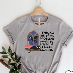 May include: A heather gray t-shirt featuring a graphic of a black cat lying on a stack of books next to a candle and a window with a silhouette of a dragon. The text reads: I THINK a lot of my PROBLEMS would be solved if I had a DRAGON.