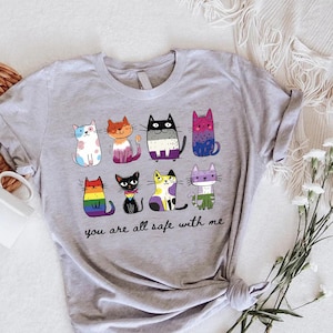 May include: A light gray t-shirt featuring a colorful illustration of ten cartoon cats with different patterns and colors. The text "you are all safe with me" is printed below the cats. The shirt is made of a soft material.