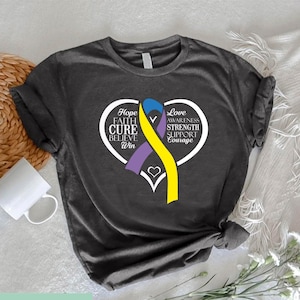 May include: Dark gray t-shirt featuring a white heart with the words "Hope, Faith, Cure, Believe, Win, Love, Awareness, Strength, Support, Courage." A purple and yellow ribbon is inside the heart. The shirt has a small logo at the bottom.