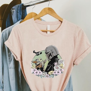 Peut inclure: Un t-shirt rose clair avec un motif d'un personnage portant un casque et une petite cr&eacute;ature, entour&eacute;s de fleurs. Le t-shirt est suspendu &agrave; un cintre en bois avec une chemise en jean bleue et un chapeau marron en arri&egrave;re-plan.