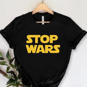 May include: Black t-shirt with the words "STOP WARS" in yellow, displayed on a wooden hanger. The text is in a bold, sans-serif font. A sprig of greenery is placed to the left of the shirt.