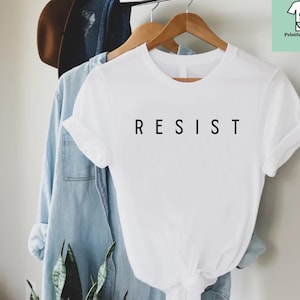 以下が含まれることがあります： 前面に黒で「RESIST」とプリントされた白いTシャツです。