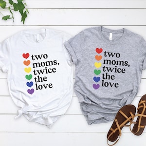 May include: Two t-shirts, one white and one gray, with the text "two moms, twice the love" in black. Each shirt has a rainbow of hearts. Brown sandals are next to the gray shirt. The shirts are on a white wooden surface.