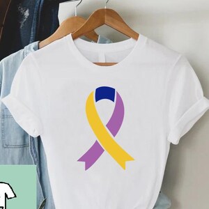 May include: White t-shirt with a colorful ribbon graphic in yellow, purple, and blue. The shirt is hanging on a wooden hanger with other clothing items. The shirt has a round neck and short sleeves.