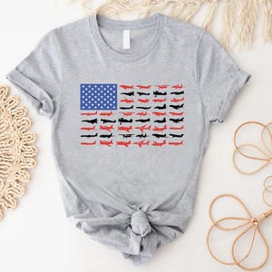 Peut inclure: T-shirt gris clair avec un motif du drapeau am&eacute;ricain compos&eacute; d'avions. Les &eacute;toiles du drapeau sont bleues et les rayures sont compos&eacute;es de silhouettes d'avions rouges et noires. Le t-shirt a des manches courtes et un n&oelig;ud nou&eacute; en bas.