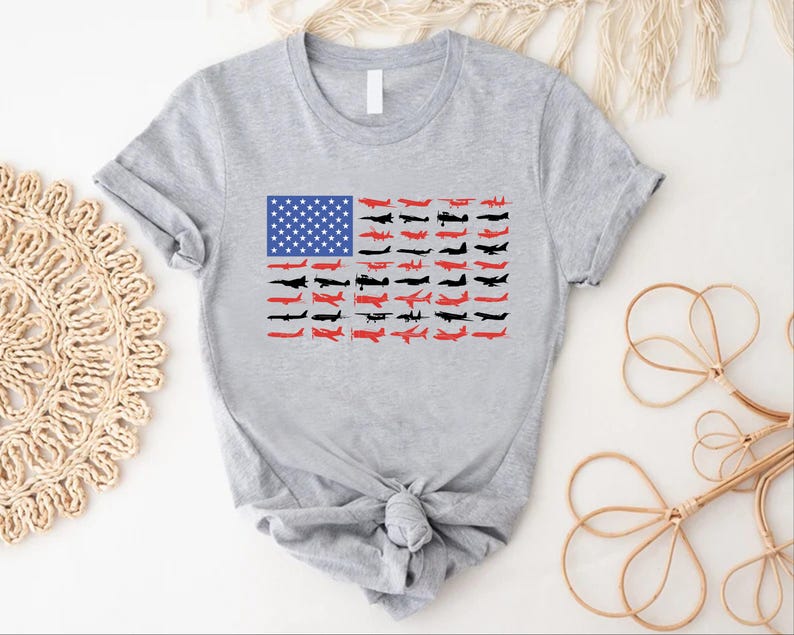Peut inclure: T-shirt gris clair avec un motif du drapeau am&eacute;ricain compos&eacute; d'avions. Les &eacute;toiles du drapeau sont bleues et les rayures sont compos&eacute;es de silhouettes d'avions rouges et noires. Le t-shirt a des manches courtes et un n&oelig;ud nou&eacute; en bas.