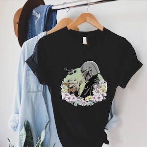 Peut inclure: T-shirt noir avec un motif de The Mandalorian et Grogu entour&eacute;s de fleurs. Le motif comprend une figurine casqu&eacute;e et une petite cr&eacute;ature adorable. Le t-shirt est suspendu &agrave; un cintre en bois avec une chemise en jean et un chapeau marron.