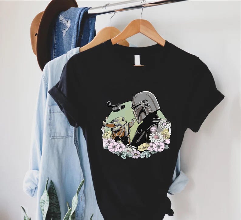 Peut inclure: T-shirt noir avec un motif de The Mandalorian et Grogu entour&eacute;s de fleurs. Le motif comprend une figurine casqu&eacute;e et une petite cr&eacute;ature adorable. Le t-shirt est suspendu &agrave; un cintre en bois avec une chemise en jean et un chapeau marron.