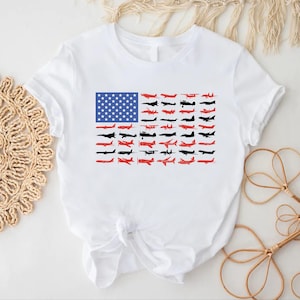Peut inclure: T-shirt blanc avec un motif du drapeau am&eacute;ricain compos&eacute; d'avions. Le motif du drapeau comprend un rectangle bleu avec des &eacute;toiles blanches et des rang&eacute;es de silhouettes d'avions rouges et noires.