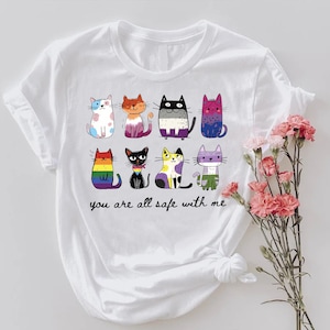 May include: White t-shirt featuring a colorful illustration of nine cartoon cats with various patterns and colors. The text "you are all safe with me" is printed below the cats. A bouquet of pink flowers is on the right.