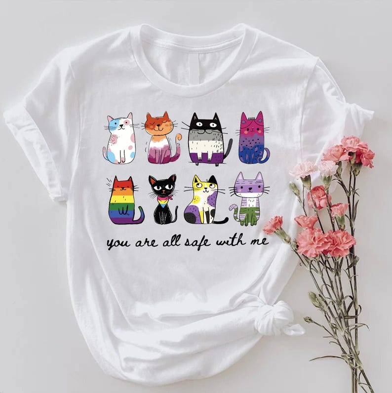 May include: White t-shirt featuring a colorful illustration of nine cartoon cats with various patterns and colors. The text "you are all safe with me" is printed below the cats. A bouquet of pink flowers is on the right.