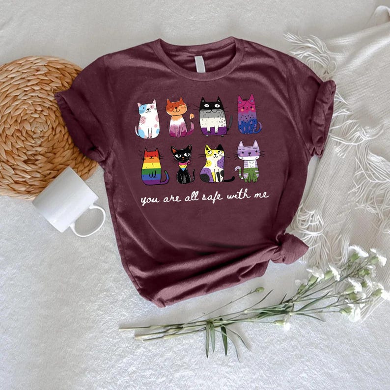 May include: Burgundy t-shirt featuring a colorful graphic of nine stylized cats, each with a unique pattern and color scheme. The text "you are all safe with me" is printed below the cats. The shirt is displayed on a white surface.