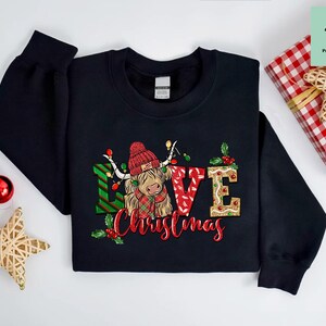 K&ouml;nnte beinhalten: Schwarzer Pullover mit einem Weihnachtsmotiv, das eine schottische Hochlandkuh mit einem Weihnachtsmann-Hut und den Worten "Love Christmas" zeigt.