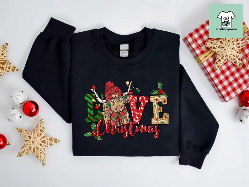 K&ouml;nnte beinhalten: Schwarzer Pullover mit einem Weihnachtsmotiv, das eine schottische Hochlandkuh mit einem Weihnachtsmann-Hut und den Worten "Love Christmas" zeigt.
