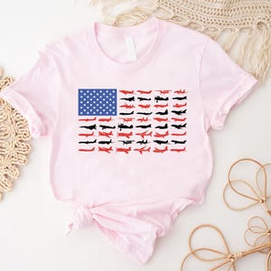 Peut inclure: Un t-shirt rose clair avec un motif du drapeau am&eacute;ricain compos&eacute; d'avions. Le champ bleu du drapeau est rempli d'&eacute;toiles blanches, et les rayures sont repr&eacute;sent&eacute;es par des silhouettes d'avions rouges et noirs. Le t-shirt est nou&eacute; &agrave; la taille.