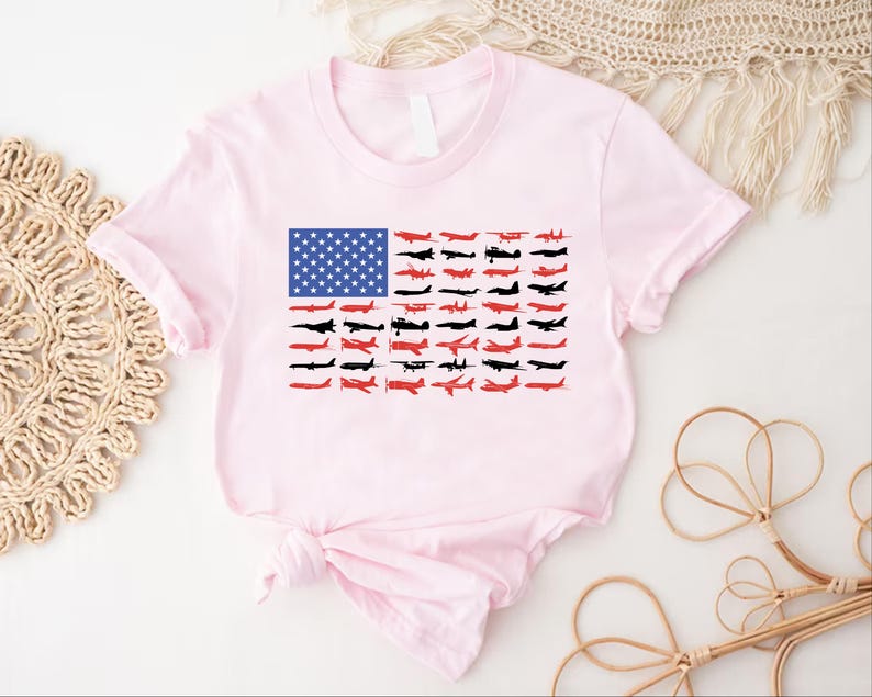 Peut inclure: Un t-shirt rose clair avec un motif du drapeau am&eacute;ricain compos&eacute; d'avions. Le champ bleu du drapeau est rempli d'&eacute;toiles blanches, et les rayures sont repr&eacute;sent&eacute;es par des silhouettes d'avions rouges et noirs. Le t-shirt est nou&eacute; &agrave; la taille.