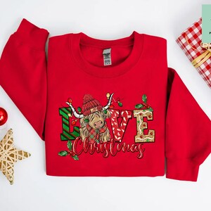 K&ouml;nnte beinhalten: Roter Weihnachtspullover mit einem festlichen Design, das eine Highland-Kuh mit einem rot-gr&uuml;nen karierten Schal und den Worten "Love Christmas" zeigt.