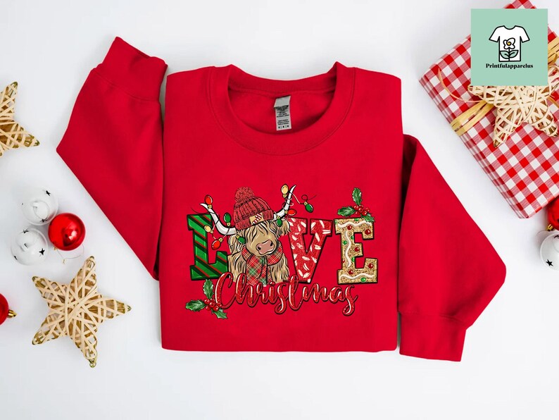K&ouml;nnte beinhalten: Roter Weihnachtspullover mit einem festlichen Design, das eine Highland-Kuh mit einem rot-gr&uuml;nen karierten Schal und den Worten "Love Christmas" zeigt.