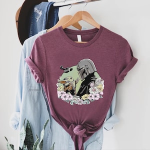Peut inclure: T-shirt bordeaux avec un motif de The Mandalorian et Grogu entour&eacute;s de fleurs. Le motif comprend une figurine casqu&eacute;e et une petite cr&eacute;ature adorable. Le t-shirt est pr&eacute;sent&eacute; sur un cintre en bois.