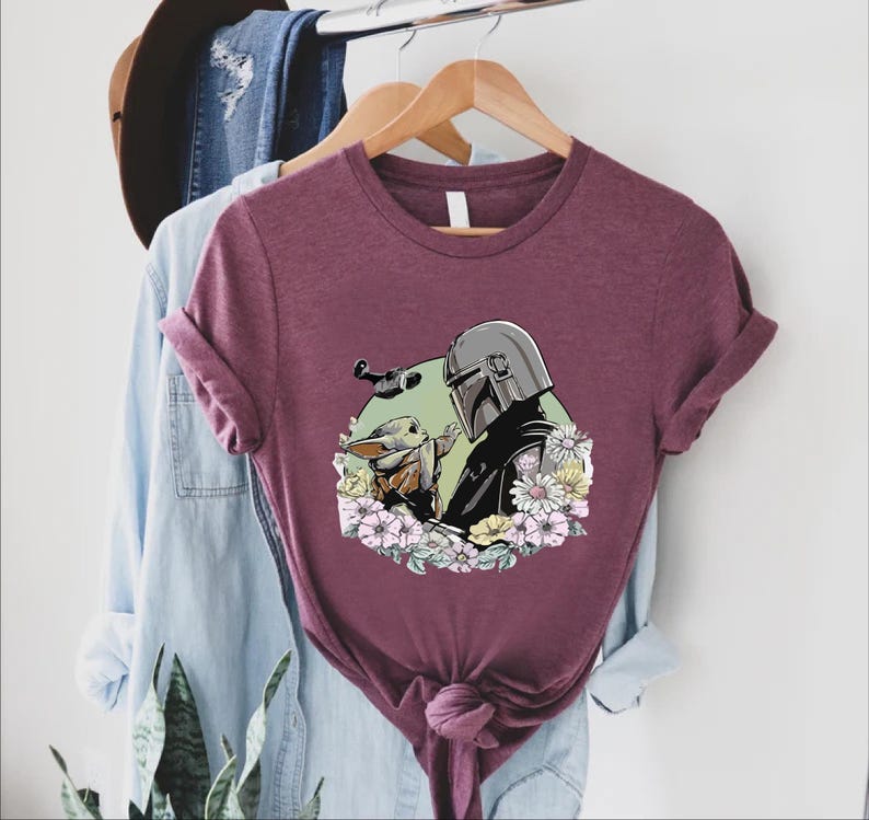 Peut inclure: T-shirt bordeaux avec un motif de The Mandalorian et Grogu entour&eacute;s de fleurs. Le motif comprend une figurine casqu&eacute;e et une petite cr&eacute;ature adorable. Le t-shirt est pr&eacute;sent&eacute; sur un cintre en bois.