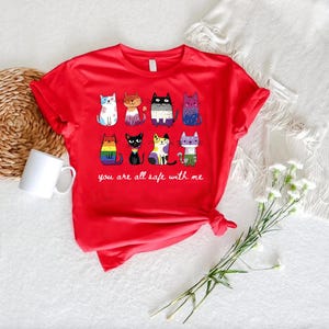 May include: A red t-shirt featuring a collection of cartoon cats in various colors and patterns. The phrase "you are all safe with me" is printed below the cats. A white mug and a bouquet of white flowers are also visible.