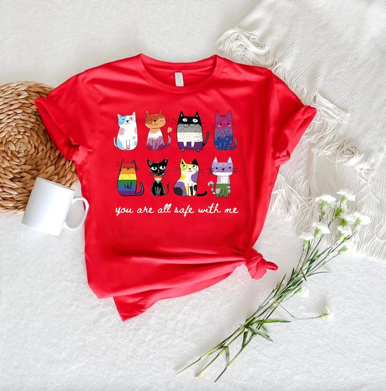May include: A red t-shirt featuring a collection of cartoon cats in various colors and patterns. The phrase "you are all safe with me" is printed below the cats. A white mug and a bouquet of white flowers are also visible.