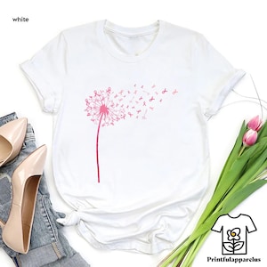 May include: White t-shirt featuring a pink dandelion design with pink ribbon accents. The dandelion stem and head are in a solid pink color, with the ribbon accents blowing in the wind. The word "white" is in the top left corner.