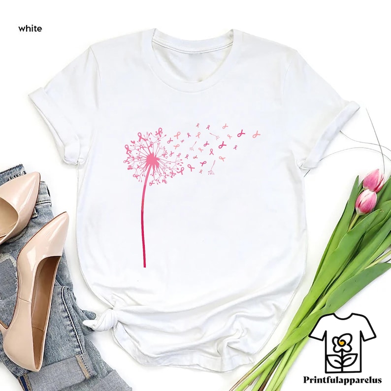 May include: White t-shirt featuring a pink dandelion design with pink ribbon accents. The dandelion stem and head are in a solid pink color, with the ribbon accents blowing in the wind. The word "white" is in the top left corner.