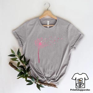 May include: A heather gray t-shirt featuring a pink dandelion design. The dandelion's stem and head are pink, with the seeds blowing away. The shirt is hanging on a wooden hanger. The text "Printfulapparelus" is in the bottom right corner.