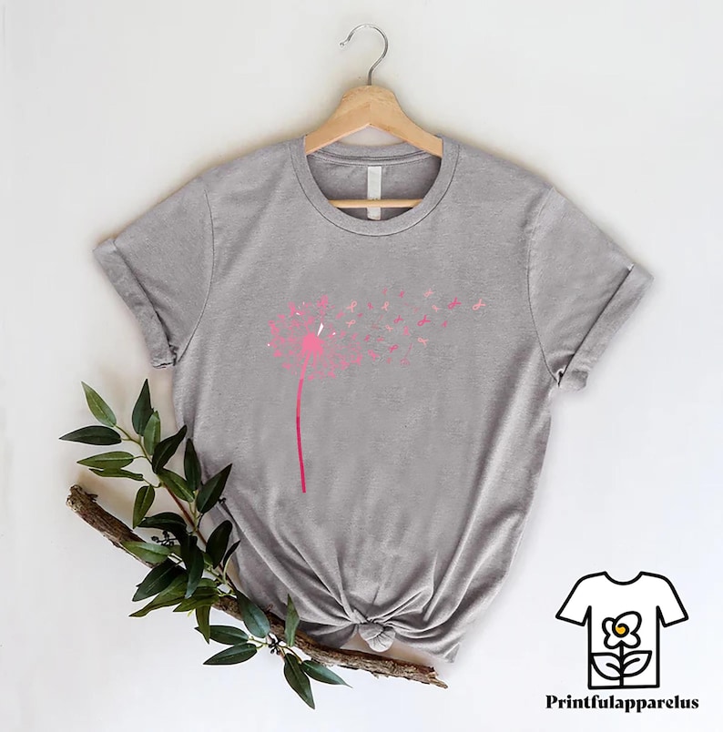 May include: A heather gray t-shirt featuring a pink dandelion design. The dandelion's stem and head are pink, with the seeds blowing away. The shirt is hanging on a wooden hanger. The text "Printfulapparelus" is in the bottom right corner.