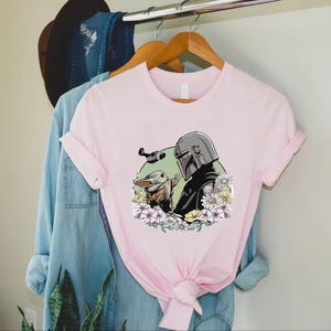 Peut inclure: T-shirt rose clair avec un motif de The Mandalorian et Grogu entour&eacute;s de fleurs. Le Mandalorien porte son casque et Grogu tend la main. Un petit vaisseau spatial vole au-dessus d'eux.