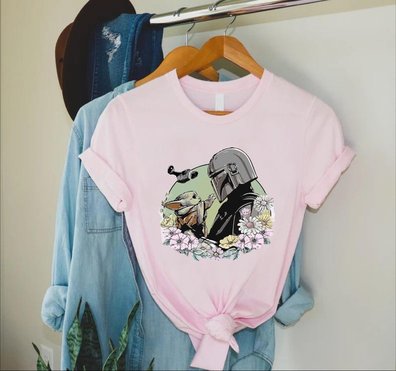 Peut inclure: T-shirt rose clair avec un motif de The Mandalorian et Grogu entour&eacute;s de fleurs. Le Mandalorien porte son casque et Grogu tend la main. Un petit vaisseau spatial vole au-dessus d'eux.
