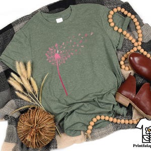 May include: A heather green t-shirt featuring a pink dandelion design with pink ribbon accents. The shirt is displayed with brown leather shoes, a wooden bead necklace, and a decorative woven basket. The Printfulapparelus logo is in the bottom right corner.