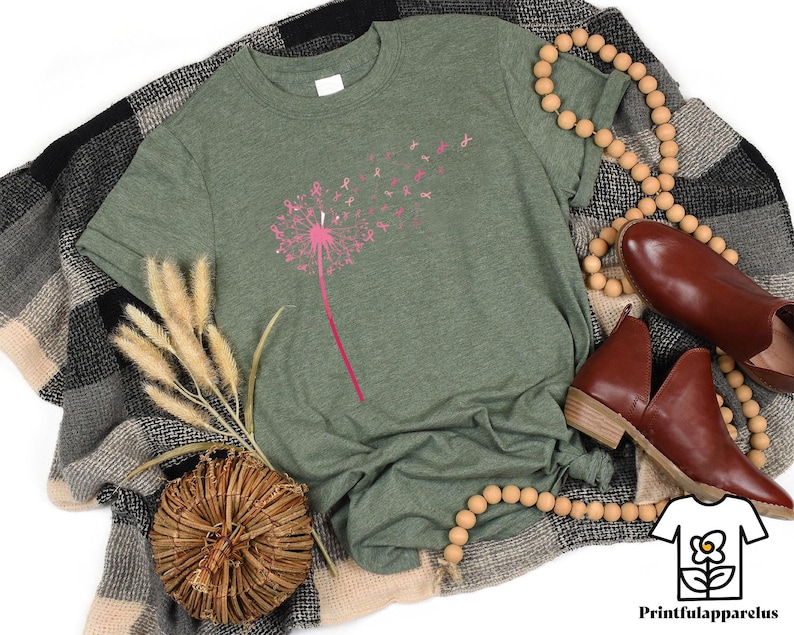 May include: A heather green t-shirt featuring a pink dandelion design with pink ribbon accents. The shirt is displayed with brown leather shoes, a wooden bead necklace, and a decorative woven basket. The Printfulapparelus logo is in the bottom right corner.