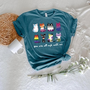 May include: Teal t-shirt featuring a colorful graphic of nine cartoon cats, each with a unique design. The text "you are all safe with me" is printed below the cats. The shirt is made of soft material.