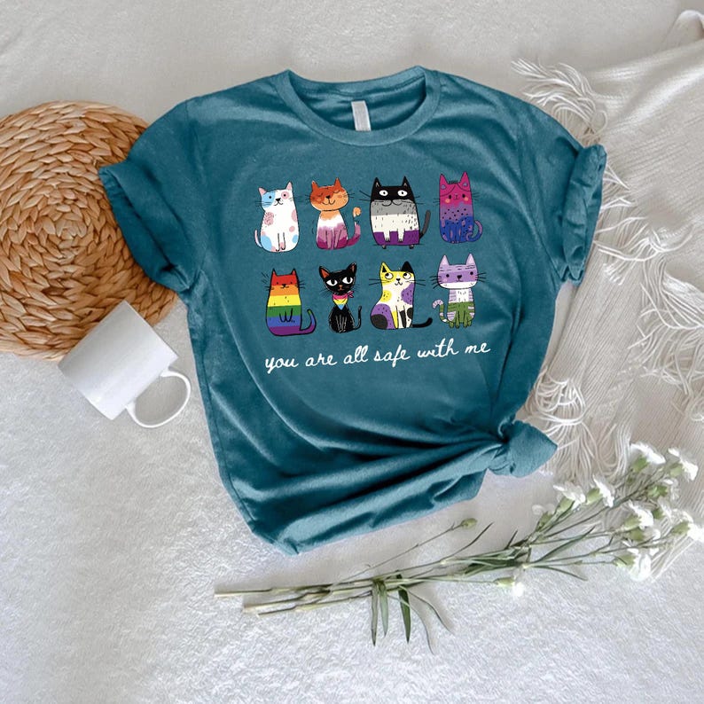 May include: Teal t-shirt featuring a colorful graphic of nine cartoon cats, each with a unique design. The text "you are all safe with me" is printed below the cats. The shirt is made of soft material.